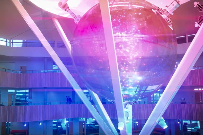 Fusion building - lab An atrium of a large building with corridors 3 stories of high in the background. From the ceiling there are some computerised special effects showing a ball with three pillars coming down from it
