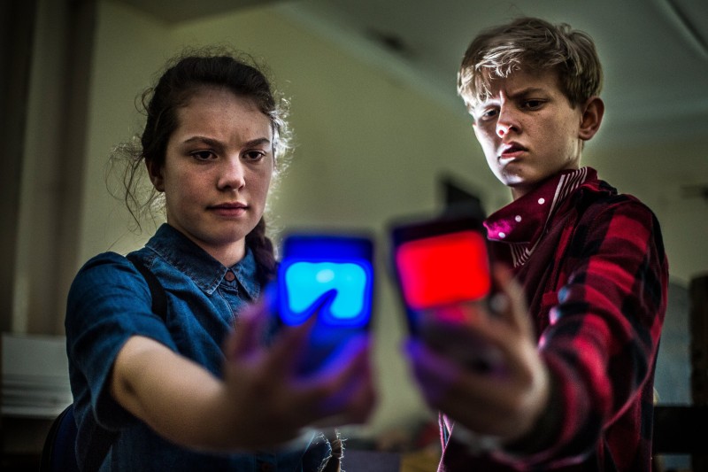 A young boy and girl holding their phones out and looking inquisitively at the screens. Her phone has a large blue light on the back of it, his has a red light