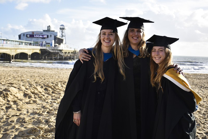 Three ladies standing on the beach in their graduation robes and hats, with Bournemouth pier in the background