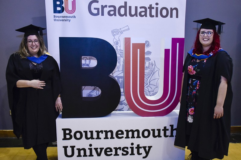 Two ladies wearing their graduation robes and hats, looking at the camera smiling, standing either side of a large banner with the BU Logo