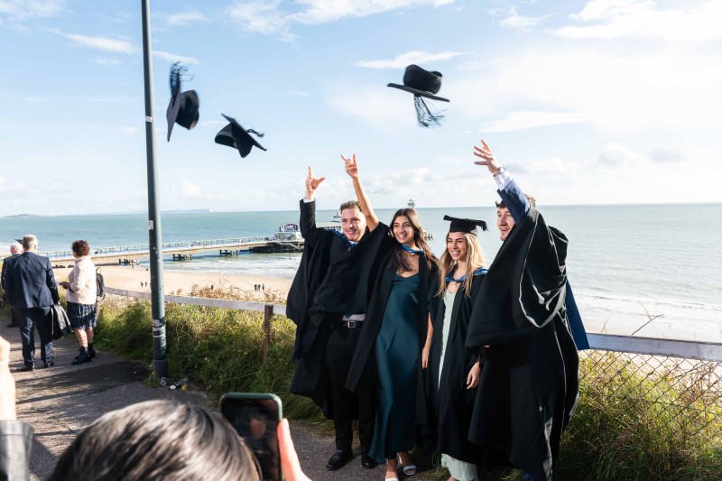 Graduates 2023 Four graduates in their gowns, throwing their hats in the air, with Bournemouth beach and pier in the background