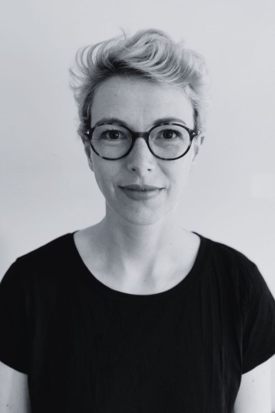 This black and white photo shows Harriet smiling to the camera. Her hair is short and styled, she wears a black t-shirt and rounded galsses