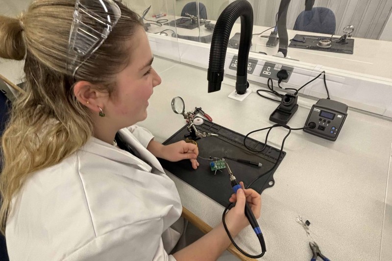 Chloe in a workshop holding an electrical device she is working on in one hand and a tool in the other