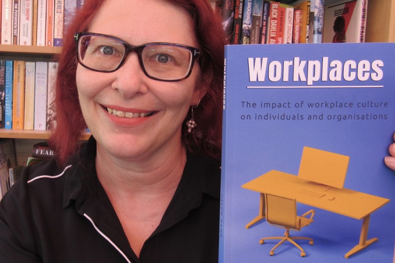 Helen Rana - head and shoulders photo in front of a bookshelf holding up a copy of her book