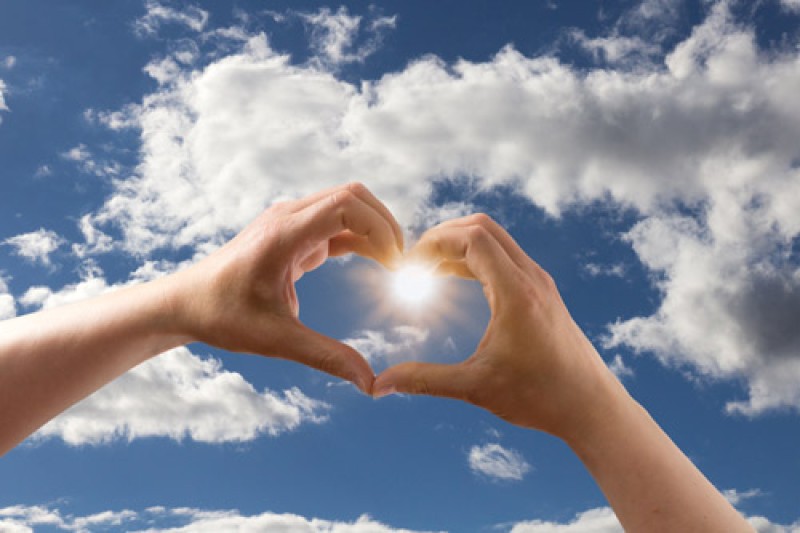 Two hands being held in a heart shape up to the sky with clouds and sunshine