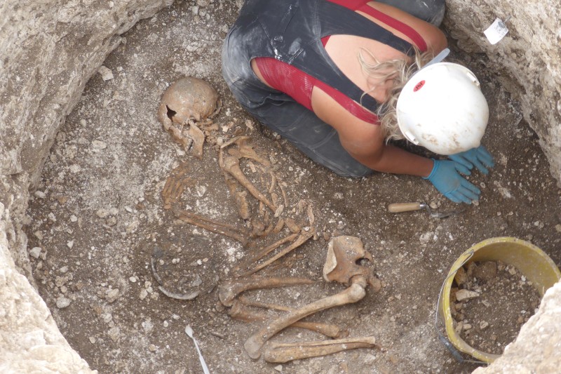 A female archaeology student, crounched in a pit, next to an old skeleton in a crouched positionn in a 