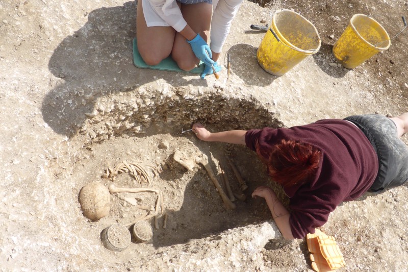Two female archaeology students looking over an old skeleton in a crounched position in a shallow pit. Two old, ceramic pots are next to the skeleton