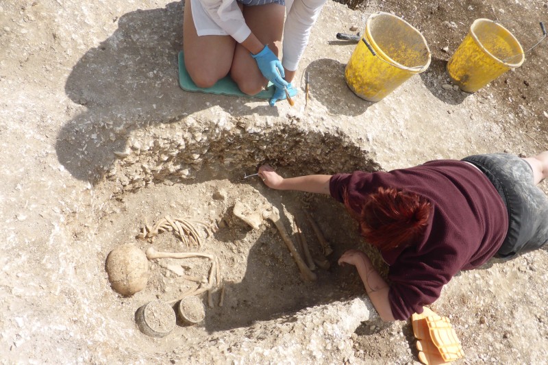 Students and Iron Age Skeleton Two female archaeology students looking over an old skeleton in a crounched position in a shallow pit. Two old, ceramic pots are next to the skeleton