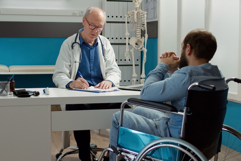 A man in a wheelchair sits in front of a desk. A doctor sits behind the desk filling out a form