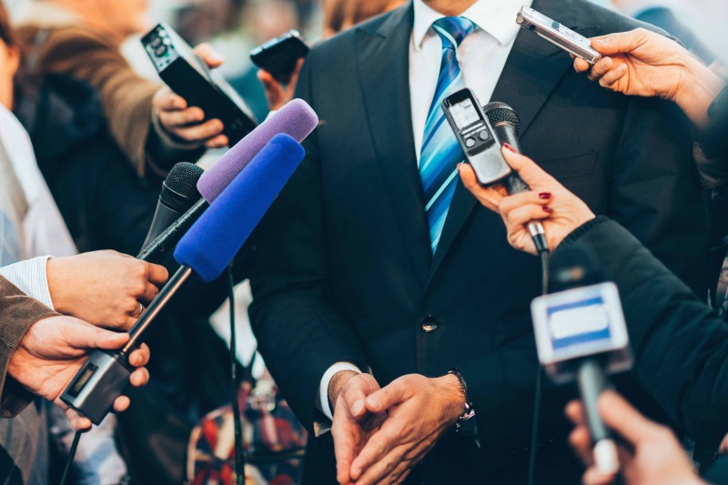 A man in a suit surrounded by people holding microphones towards him