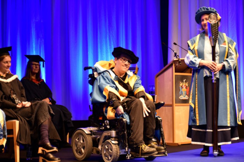 James Rose in his wheelchair, wearing ceremonial robes, holding his certificate. He is sitting either side of two ladies, also also in ceremonial robes