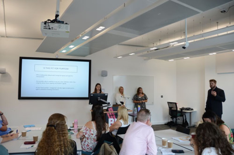 three female students and a male lecturer at the front of a room talking to people sitting on tables