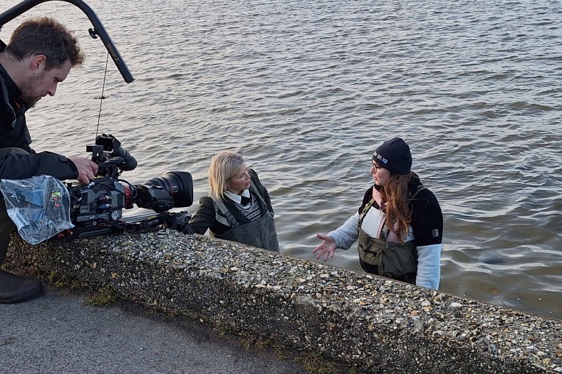 Jess Bone and Presenter Ellie Harrison wearing waders in Poole Harbour, a cameraman stands on the harbour wall, filming them.