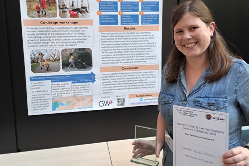 Malika holding her certificate and trophy in front of a poster depicting her research