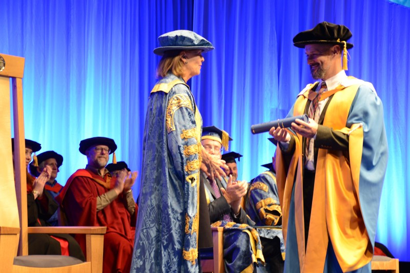 Max McMurdo Max Murdo standing on the stage in his ceremonial graduation robes, receiving a scroll from a lady also in ceremonial robes