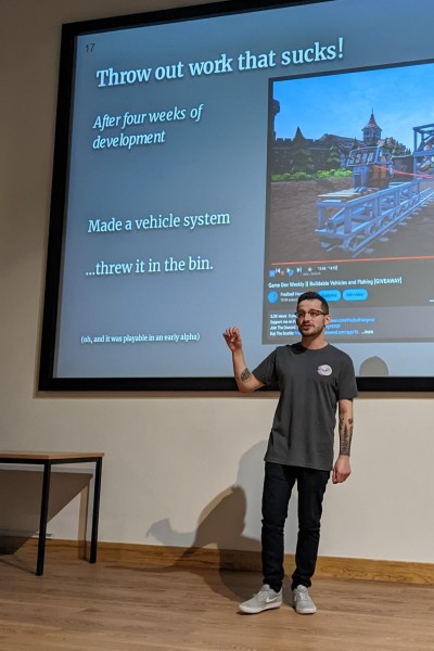 A man talking whilst standing in front of a projector screen, on the screen the title says "Throw out work that sucks, after four weeks of development, made a vehicle system, threw it in the bin"