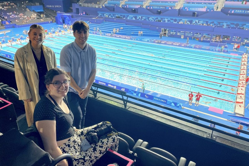 Two female students and one male students in the swimming arena press box, with the pool in the background