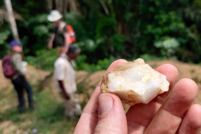 a white stone being held up to the camera, researchers working on the site in the background