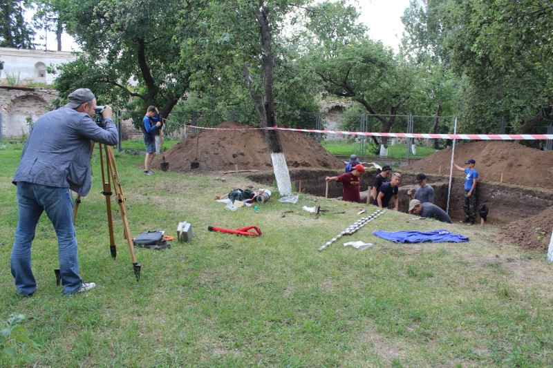 A man surveying an archaeological pit, a group of young archaeologists are inside the pit excavating 