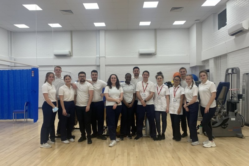 A group of fourteen people in physiotherapist uniform, standing in a row, looking at the camera, in a gymnasium