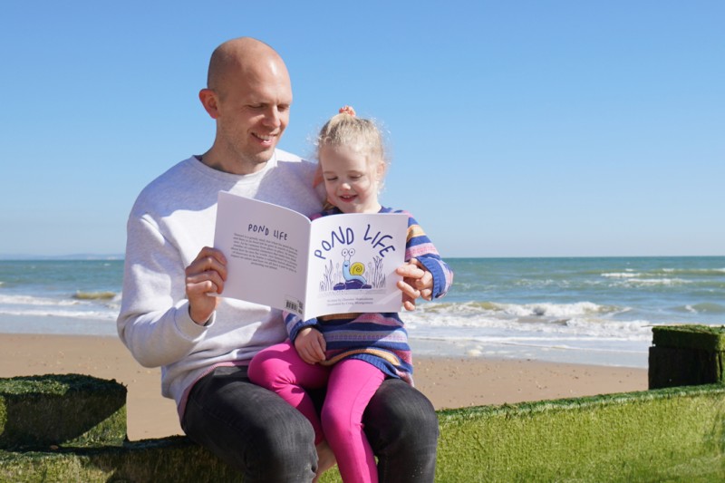 Pond life author sitting on the beach reading the book to his young daughter