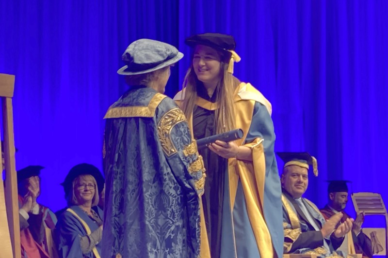 Poppy Cleall in her graduation gown and cap receiving a scrolled certificate from a lady also in ceremonial gowns