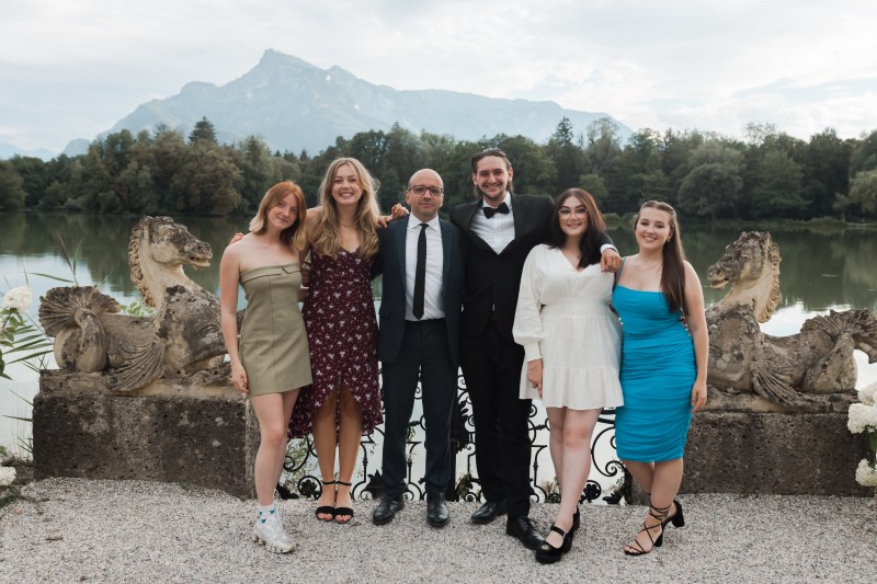 four women and two men in a line look towards the camera, a lake is behind them with some woods on the other side and mountains are in the background