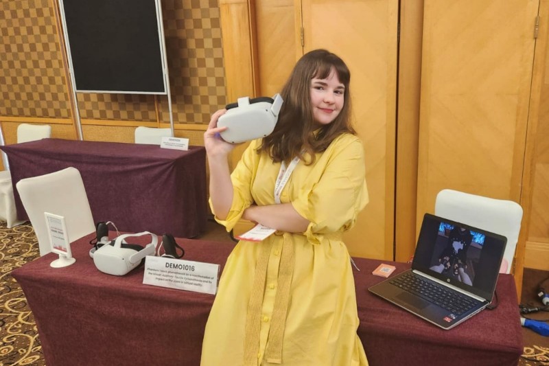 Sasha leaning against her desk, holding a Virtual Reality headset. Computer equipment is lined across the desk