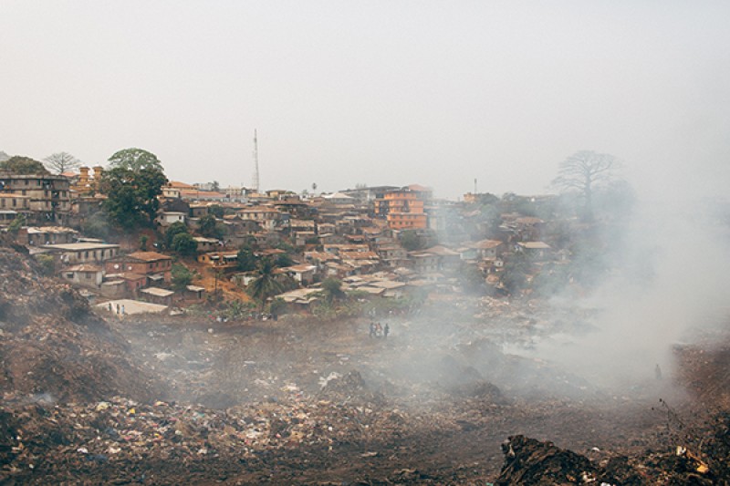 People walking next to a vast landfill site in Freetown, Sierra Leone, with smoke rising from the site