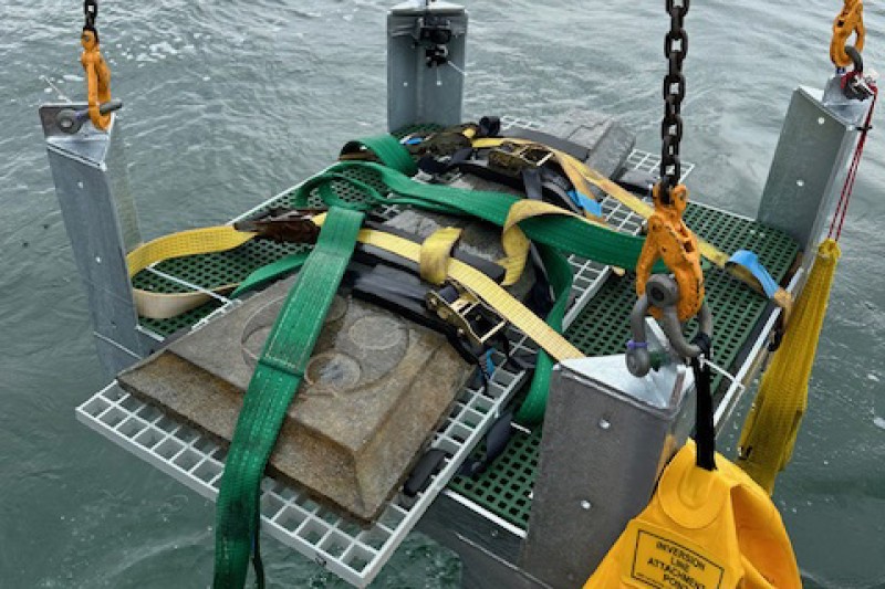 A grave stone lying on a plastic tray with chains attached to each corner, being lifted out of the sea