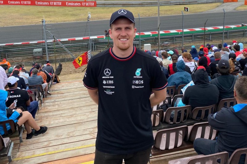 I man facing the camera in an aisle in a sports stadium with rows of seats with fans sitting on them either side of him. Behind him is a motor sport racing track