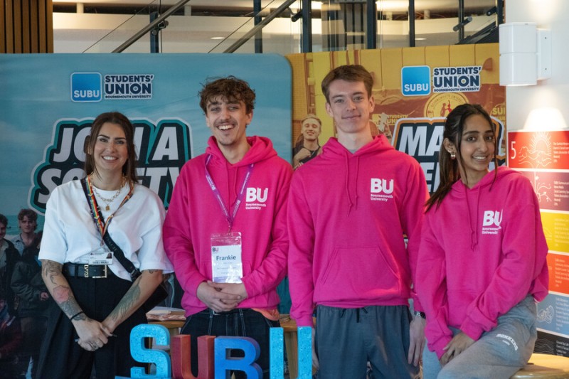 Three Bournemouth University student ambassadors wearing bright pink hoodies stood in front of Students' Union Bournemouth University (SUBU) banners in the Student Centre