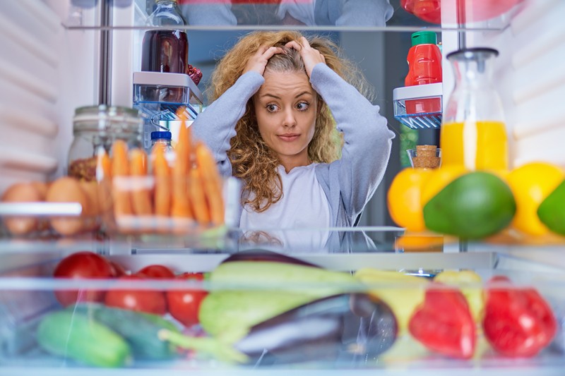 View from inside the fridge containing lots of vegetables. In the background is a lady facing towards the camera with a confused expression on her face. 