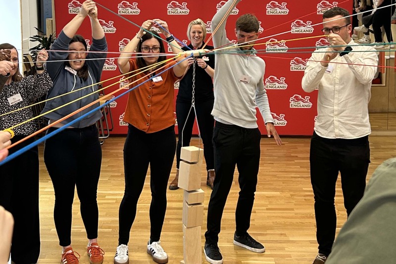 A team of people each holding on to pieces of string. The strings are attached to wooden blocks which they are trying to stack into a tower