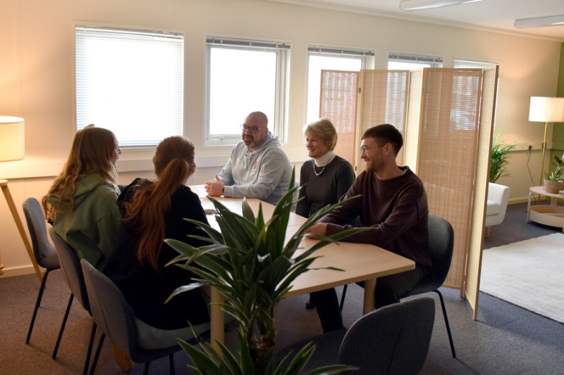 Six people stand around a table at the University Retreat on Talbot Campus. The decor is neutral tones with plants dotted around the room