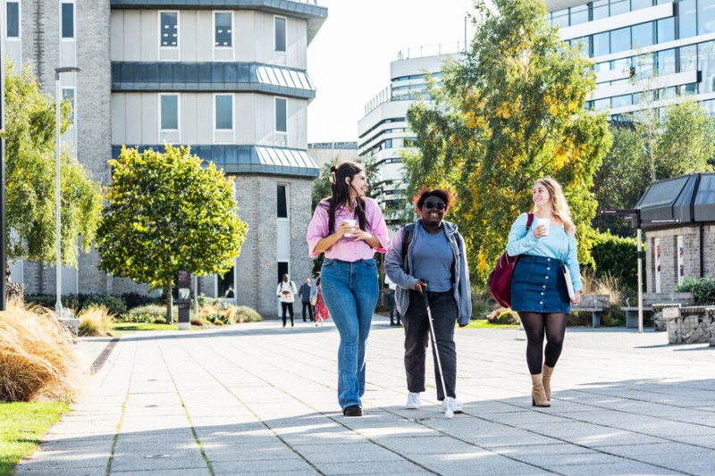 Three students walking on campus with buildings and trees in background