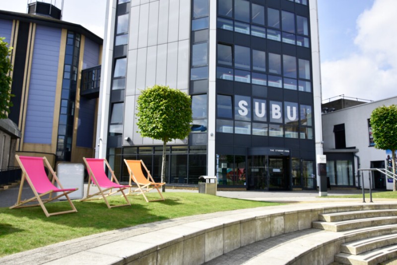 An exterior shot of the Student Centre. A modern buildings with solar panels and lots of windows. In the foreground are 3 deckchairs