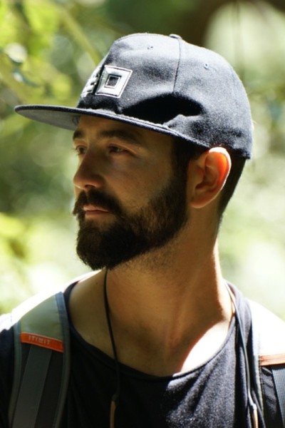 Alfonso is pictured in the outdoors, he is central in this image wearing a baseball cap, black t-shirt and a rucksack. His head is turned to the left.