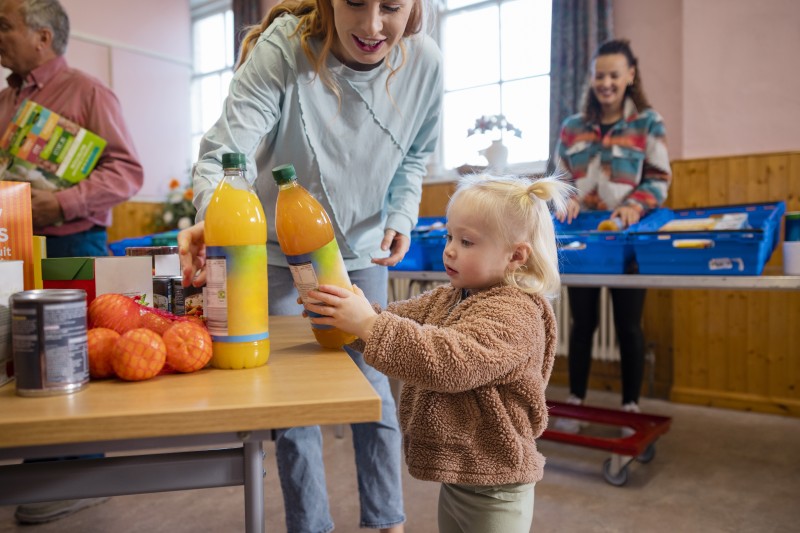 Market image mother and daughter picking up food together at a food bank. It is being run by volunteers at a community church in the North East of England. The focus is on a young girl is holding a bottle of juice.