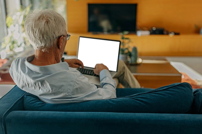 View over the shoulder of an older man sitting on a sofa looking at his tablet