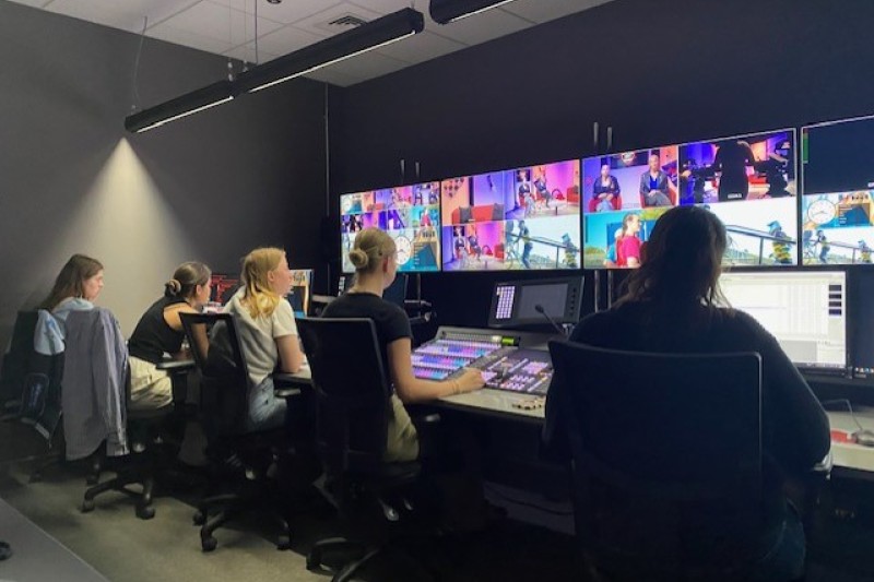 Four students sit in front of a wall of TV screens, each showing multiple broadcasts