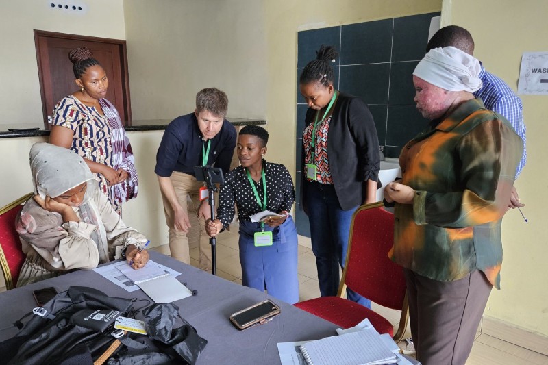 A video workshop - one student is sitting at a desk reading. Another female student is filming her with a mobile phone, facilitators are standing nearby